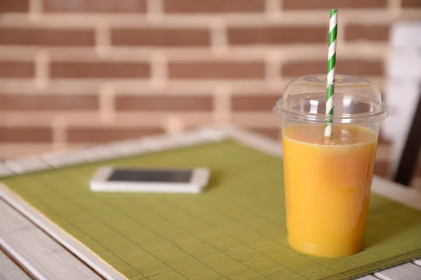 Jugo de naranja en taza cerrada de comida rápida con tubo y teléfono móvil en mesa de madera y fondo de pared de ladrillo —  Fotos de Stock