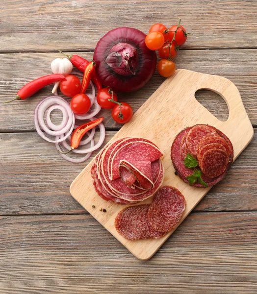 Salami en rodajas con chile, tomates cherry, cebolla y especias sobre tabla de cortar y fondo de mesa de madera — Foto de Stock