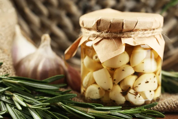 Ingeblikte knoflook in glazen pot en rieten mat en rozemarijn takken, op houten achtergrond — Stockfoto