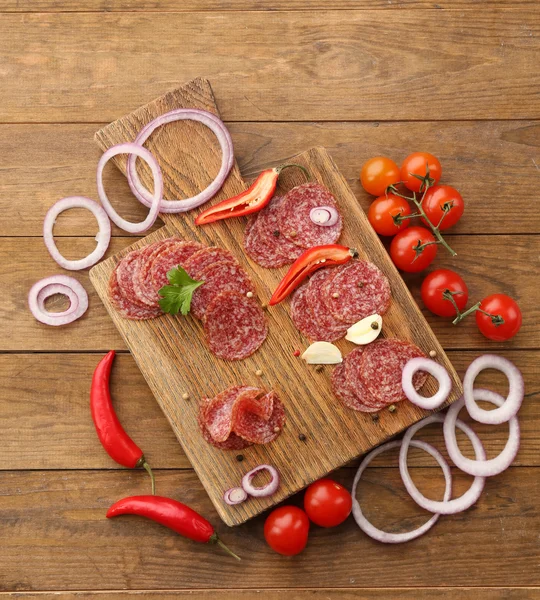 Rodajas de salami con especias y verduras sobre tabla de cortar y fondo de mesa de madera — Foto de Stock