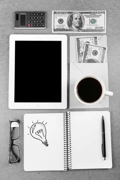 Symbol of idea as light bulb in notebook with crumpled paper, tablet, money and cup of coffee on wooden desk background — Stock Photo, Image