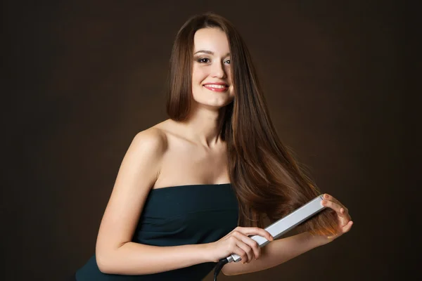 Beautiful young woman with long hair using hair straighteners on dark brown background — Stock Photo, Image