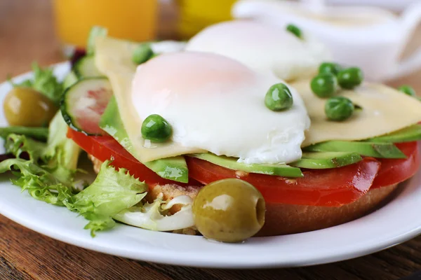 Sandwich with poached eggs, cheese and vegetables on plate on wooden background — Stock Photo, Image