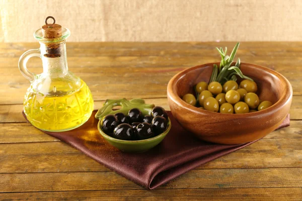 Sammansättning av två runda skålar med gröna och svarta oliver nära oljan kan på siden tyg, på rustika träbord, på säckväv bakgrund — Stockfoto