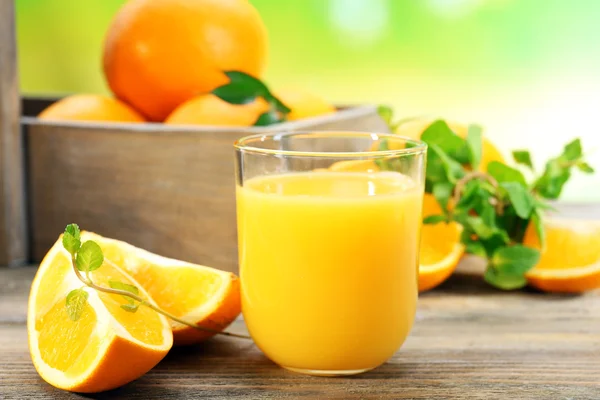 Glass of orange juice with crate of oranges and slices on wooden table and bright background — Stock Photo, Image