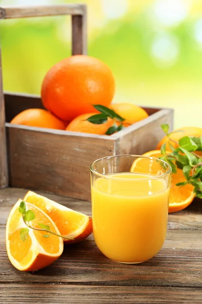 Copo de suco de laranja com caixa de laranjas e fatias na mesa de madeira e fundo brilhante — Fotografia de Stock