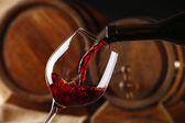 Einschenken von Rotwein aus der Flasche in Glas mit hölzernen Weinfässern auf Hintergrund