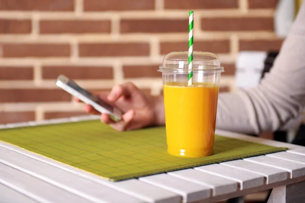 Jus d'orange in fastfood gesloten cup met buis en mobiele telefoon op houten tafel en bakstenen muur achtergrond — Stockfoto