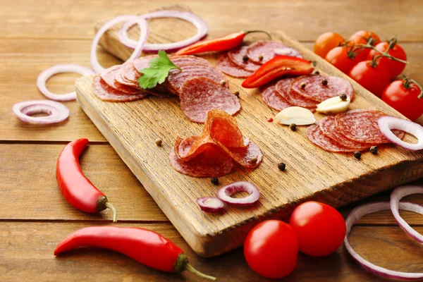 Rodajas de salami con especias y verduras sobre tabla de cortar y fondo de mesa de madera — Foto de Stock