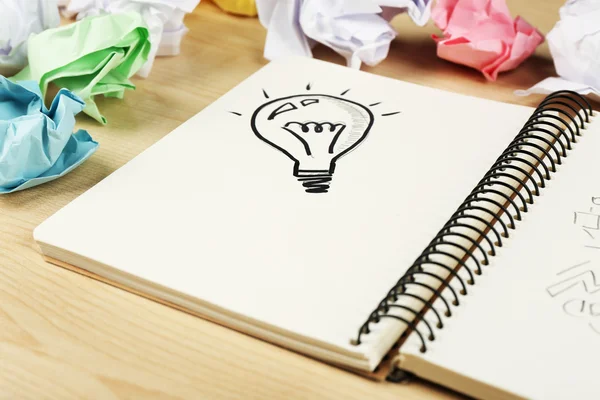 Symbol der Idee als Glühbirne im Notizbuch mit zerknittertem Papier auf hölzernem Schreibtischhintergrund — Stockfoto