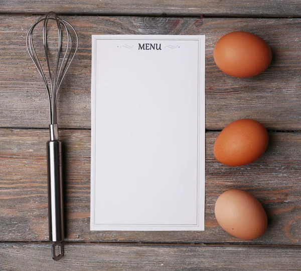 Menu sheet of paper with egg whisk and eggs on rustic wooden surface background — Stock Photo, Image