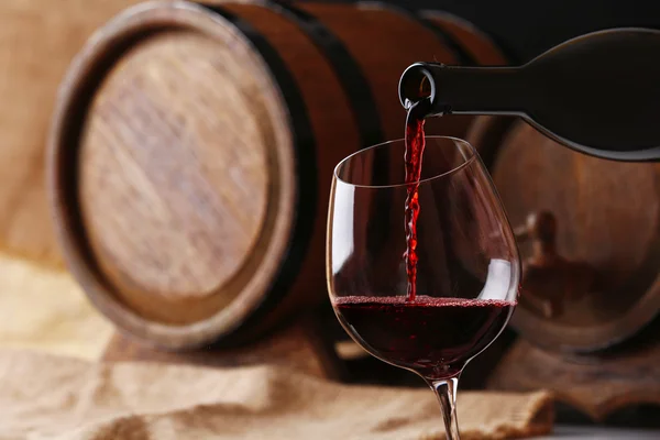 Pouring red wine from bottle into glass with wooden wine casks on background — Stock Photo, Image