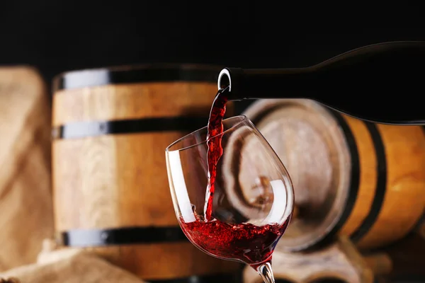 Einschenken von Rotwein aus der Flasche in Glas mit hölzernen Weinfässern auf Hintergrund — Stockfoto