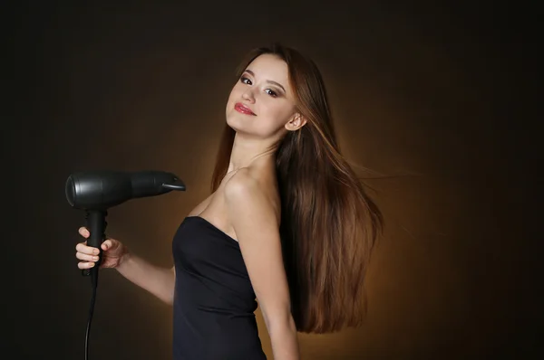 Güzel genç kadın uzun saçlı saç kurutma makinesi koyu kahverengi zemin üzerinde tutan — Stok fotoğraf