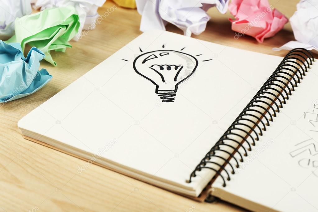 Symbol of idea as light bulb in notebook with crumpled paper on wooden desk background