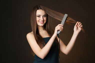 Uzun saçlı güzel bir kadın koyu kahverengi arka planda saç düzleştirici kullanıyor.