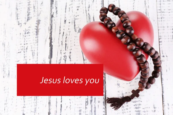Heart with rosary beads on wooden background and text Jesus loves you
