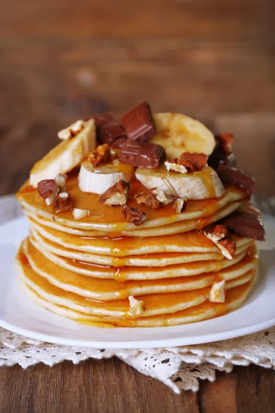 Стопка вкусных блинчиков с шоколадом, медом, орехами и ломтиками банана на тарелке и салфеткой на деревянном фоне — стоковое фото