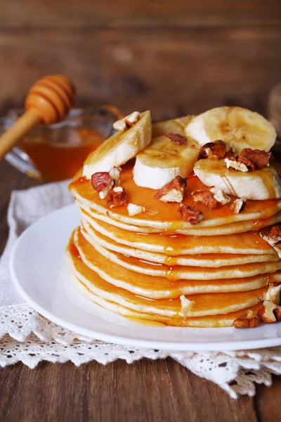 Pilha de deliciosas panquecas com chocolate, mel, nozes e fatias de banana no prato e guardanapo no fundo de madeira — Fotografia de Stock