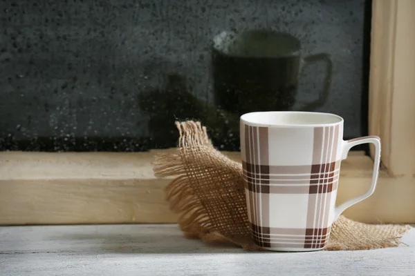 Cup of hot drink with napkin on windowsill on rain background — Stock Photo, Image