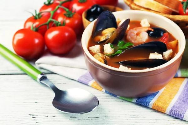 Lekkere soep met garnalen, mosselen, tomaten en zwarte olijven in kom op houten achtergrond — Stockfoto