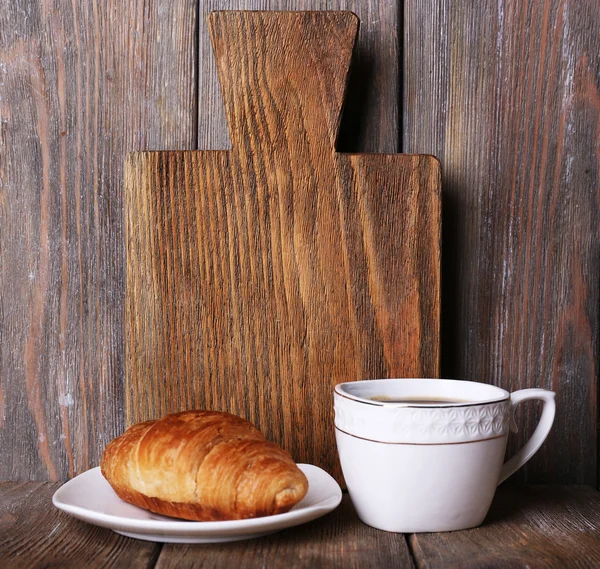 Доска для резки с чашкой кофе и круассаном на фоне деревянных досок — стоковое фото