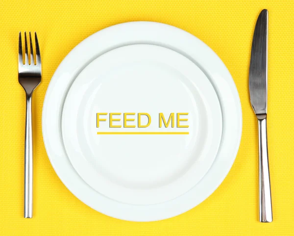 Plate with text Feed Me, fork and knife on tablecloth background — Stock Photo, Image