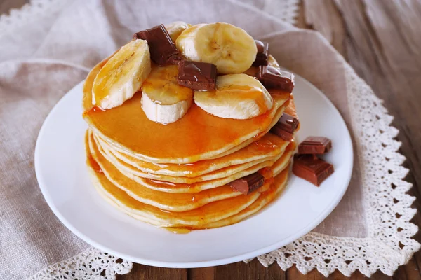 Pilha de deliciosas panquecas com chocolate, mel e fatias de banana no prato e guardanapo no fundo da mesa de madeira — Fotografia de Stock