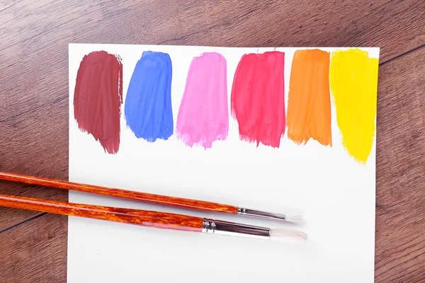 Colorful paint strokes with brush on white sheet of paper on wooden table background — Stock Photo, Image