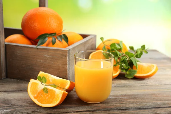 Glass of orange juice with crate of oranges and slices on wooden table and bright background — Stock Photo, Image