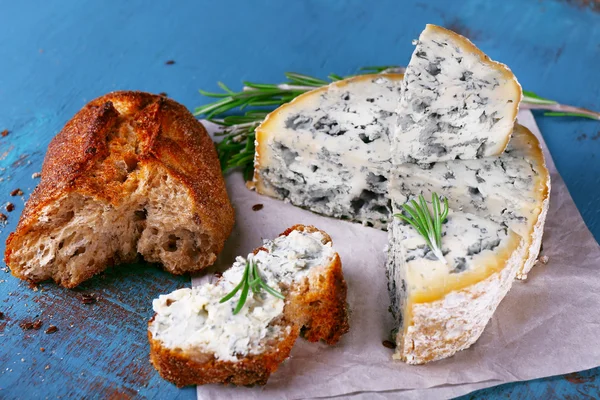 Blue cheese with sprigs of rosemary and bread on sheet of paper and color wooden table background — Stock Photo, Image