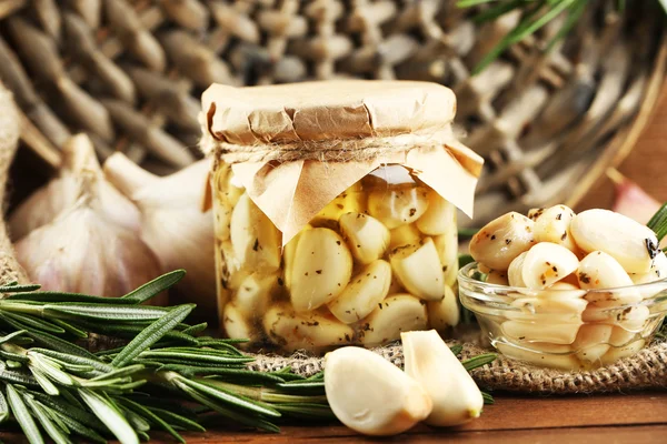 Ingeblikte knoflook in glazen pot en rieten mat en rozemarijn takken, op houten achtergrond — Stockfoto