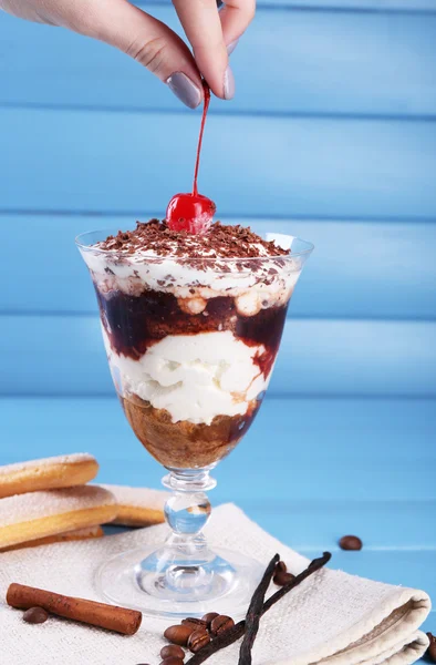 Mão segurando cereja acima saborosa sobremesa tiramisu em vidro, na cor de fundo de madeira — Fotografia de Stock