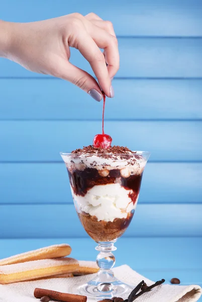 Mano che tiene ciliegia sopra gustoso dessert tiramisù in vetro, su sfondo di legno a colori — Foto Stock
