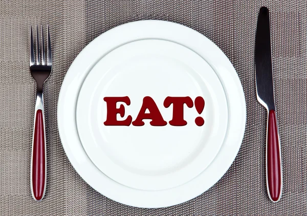 Plate with word EAT on it, fork and knife on tablecloth background — Stock Photo, Image
