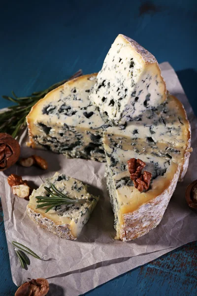 Blue cheese with sprigs of rosemary and nuts on sheet of paper and color wooden table background — Stock Photo, Image