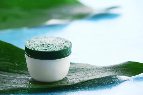 Cosmetic cream with water drops on green leaves on light background