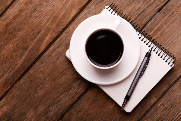 Φλιτζάνι καφέ στο πιατάκι με σημειωματάριο και στυλό σε ξύλινο τραπέζι φόντο — Φωτογραφία Αρχείου