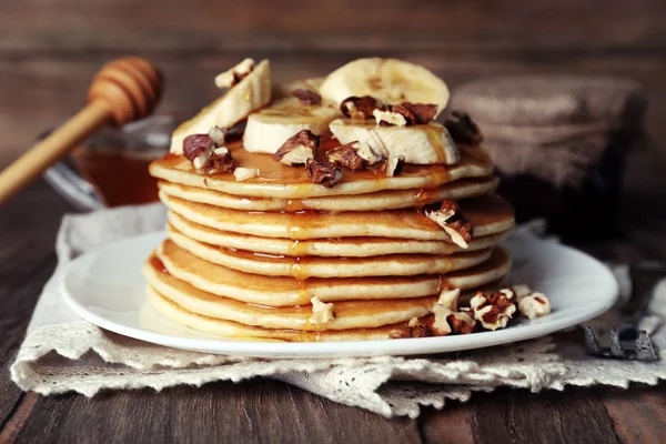 Stack of delicious pancakes with chocolate, honey, nuts and slices of banana on plate and napkin on wooden background — Stock Photo, Image