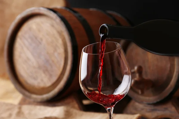 Pouring red wine from bottle into glass with wooden wine casks on background — Stock Photo, Image