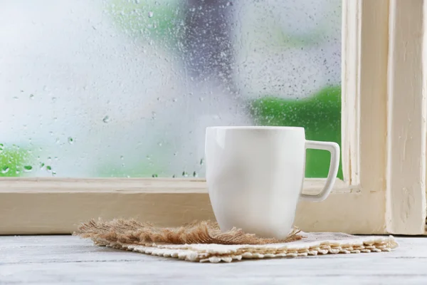 Cup of hot drink with napkin on windowsill on rain background — Stock Photo, Image