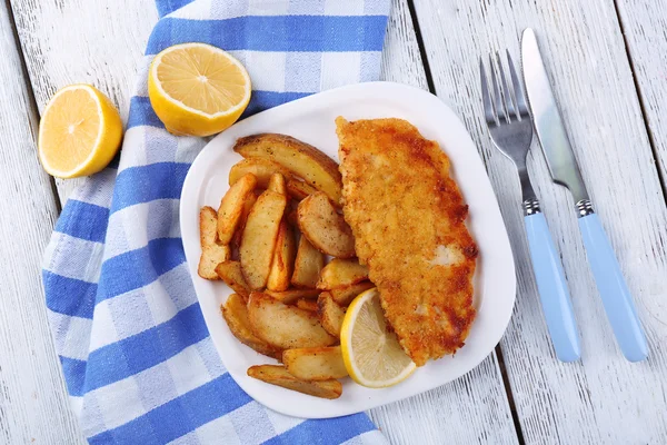 Breaded fried fish fillet and potatoes with sliced lemon on plate with napkin on color wooden planks background