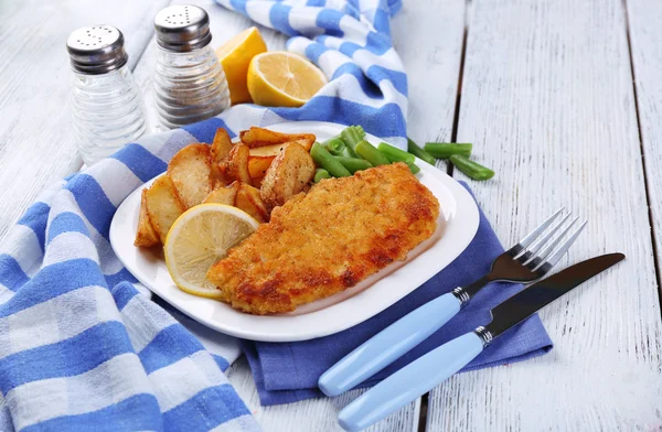 Breaded fried fish fillet and potatoes with asparagus and sliced lemon on plate with napkin on color wooden planks background — Stock Photo, Image