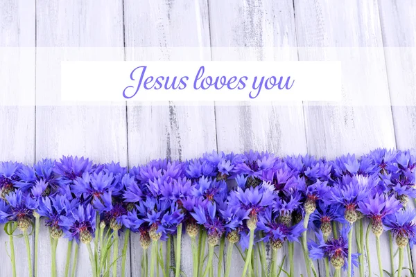 Beautiful cornflowers and text Jesus loves you on wooden background — Stock Photo, Image