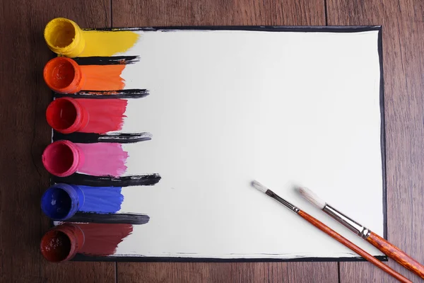 Pinturas coloridas con pincel y latas de pintura sobre papel blanco sobre fondo de mesa de madera — Foto de Stock