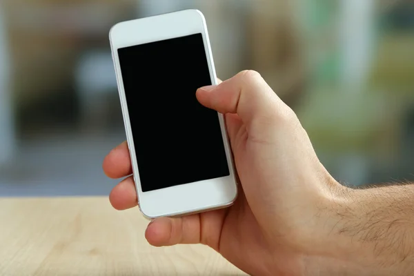 Mano que sostiene el teléfono móvil inteligente en la mesa de madera y el fondo borroso claro — Foto de Stock