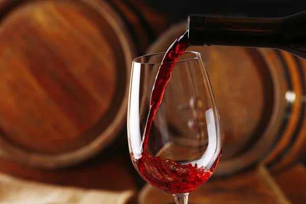 Наливание красного вина из бутылки в бокал с деревянными бочками вина на заднем плане — стоковое фото