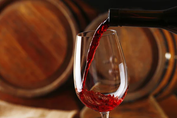 Pouring red wine from bottle into glass with wooden wine casks on background — Stock Photo, Image