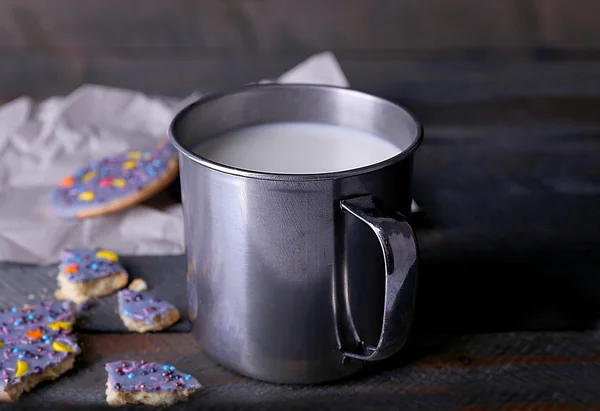 Crumbled glazed cookies on sheet of paper with metal mug of milk on rustic wooden planks background — Stock Photo, Image