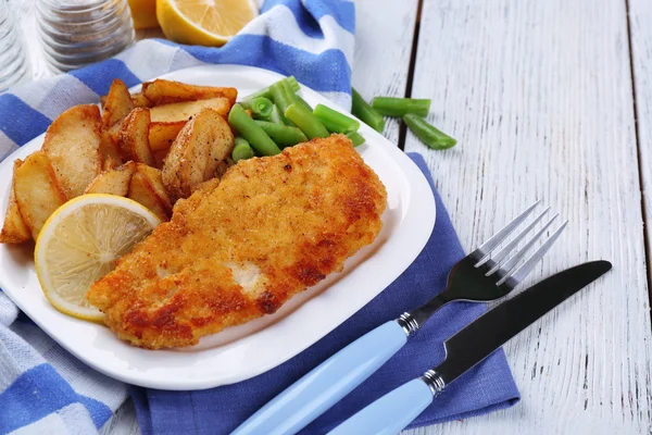 Filetto di pesce fritto impanato e patate con asparagi e limone affettato sul piatto con tovagliolo su sfondo di tavole di legno di colore — Foto Stock